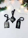 Load image into Gallery viewer, Three black holiday gift tags with hanging velvet ribbons are arranged on a white surface with Christmas greenery and warm string lights. The tags feature rhinestone-embellished designs of a reindeer, a snowflake, and a nutcracker.