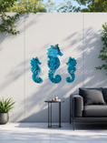 Load image into Gallery viewer, Three teal acrylic seahorses mounted with gold colored standoffs, projecting vibrant turquoise light and dancing reflections onto a patio wall.