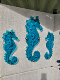 Load image into Gallery viewer, Three vivid tranparent teal acrylic seahorses with custom engraved pattern mounted with gold toned standoffs