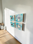 Charger l'image dans la visionneuse de la galerie, Three small square wall art panels with bird illustrations and turquoise watercolor-style backgrounds, mounted on a white wall in a sunlit room with a light wood floor and small white bottles nearby.