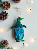 Load image into Gallery viewer, Decorative penguin resin ornament with a gradient of blue and green on a white background with pine cones and string lights.