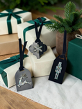 Load image into Gallery viewer, Three black holiday gift tags with silver-toned metal ornaments hang around wrapped gift boxes. The tags feature a reindeer, a snowflake, and a nutcracker design, styled with beige and green gift wrap and evergreen branches.
