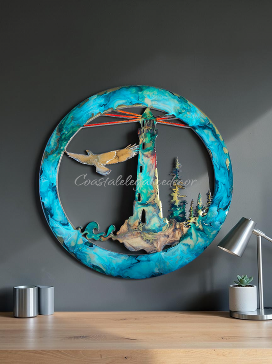 A circular 3D metal wall art piece featuring a lighthouse, trees, a bird, and abstract blue marbled borders, displayed on a wooden table against a dark gray wall with a lamp and small containers nearby.