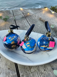Charger l'image dans la visionneuse de la galerie, Three spherical blue and white marbled ornaments with gold caps and black ribbons, displayed on a white marble tray on a weathered wooden table with the ocean and string lights in the background.
