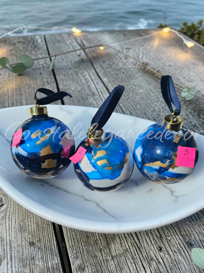 Three spherical blue and white marbled ornaments with gold caps and black ribbons, displayed on a white marble tray on a weathered wooden table with the ocean and string lights in the background.
