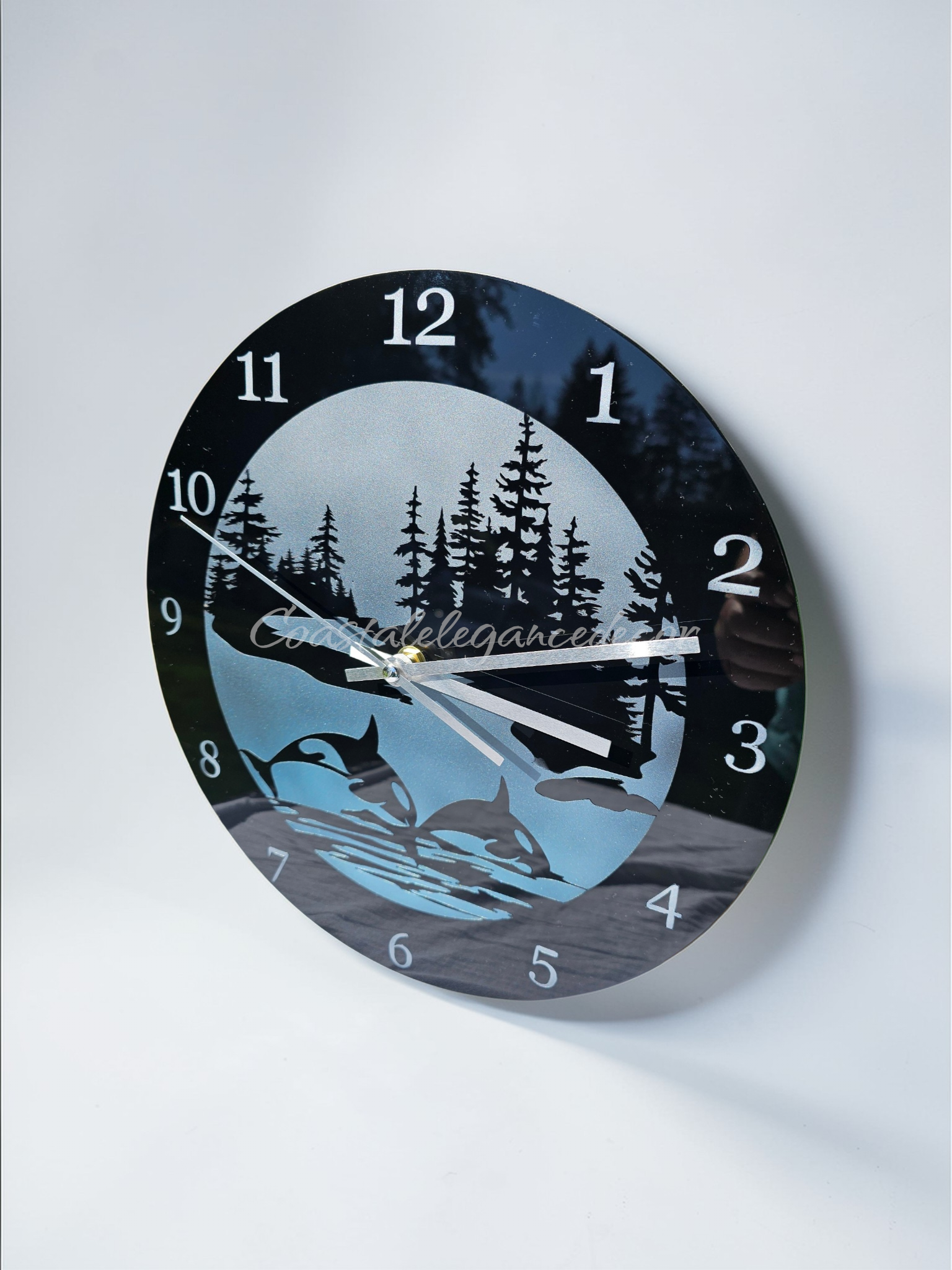 11.5 inch round acrylic wall clock featuring a hand-painted metallic blue and silver gradient side view. The design shows black silhouettes of two breaching orcas and a Pacific Northwest fir tree forest. Silver numerals and silver hands are displayed on the shimmering face.