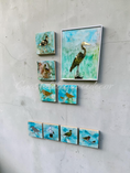 Charger l'image dans la visionneuse de la galerie, A set of bird-themed wall art pieces displayed on a textured light gray wall. The collection includes one large square canvas ink and resin of a mirrored heron and five smaller square crafle panels showing different birds, all featuring a blue and green alcohol ink background with gold mirror and silver mirror accents.
