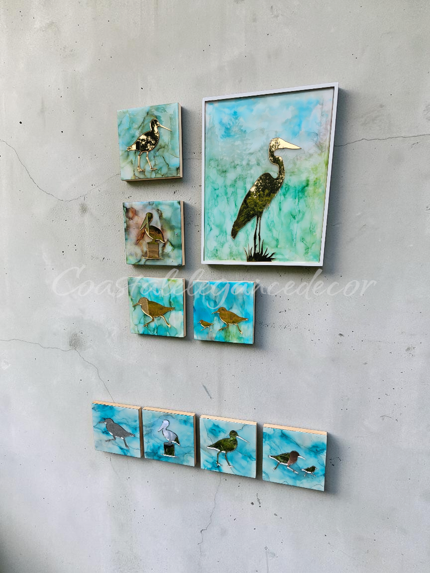 A set of bird-themed wall art pieces displayed on a textured light gray wall. The collection includes one large square canvas ink and resin of a mirrored heron and five smaller square crafle panels showing different birds, all featuring a blue and green alcohol ink background with gold mirror and silver mirror accents.