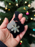 Load image into Gallery viewer, A hand holds a black Christmas ornament featuring a silver snowflake with iridescent blue and green rhinestone accents, with a blurred Christmas tree in the background.
