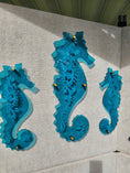 Load image into Gallery viewer, Three vivid tranparent teal acrylic seahorses with custom engraved pattern mounted with gold toned standoffs 
