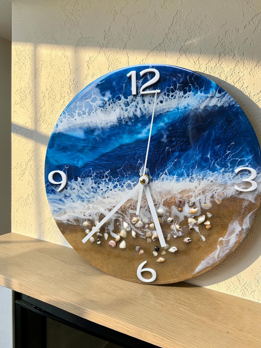 Round beach-themed wall clock with a blue ocean wave design, white foam, tan sand, and embedded seashells, shown mounted on a beige wall above a light wood surface.