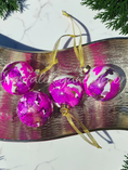 Charger l'image dans la visionneuse de la galerie, Four round glass Christmas ornaments with a vibrant magenta and white marbled pattern, hanging from gold ribbons on a metallic gold tray, framed by green pine branches on a white marble surface.
