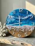 Load image into Gallery viewer, A round beach-themed wall clock with a blue ocean wave design and seashells embedded in the lower half, displayed on a rustic wooden shelf next to a white coral decoration.
