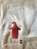 Load image into Gallery viewer, A penguin-shaped ornament or charm with a red glittery body and a silver hanging loop, placed on a light beige fabric background. A small black label with the number 4 is near the bottom.
