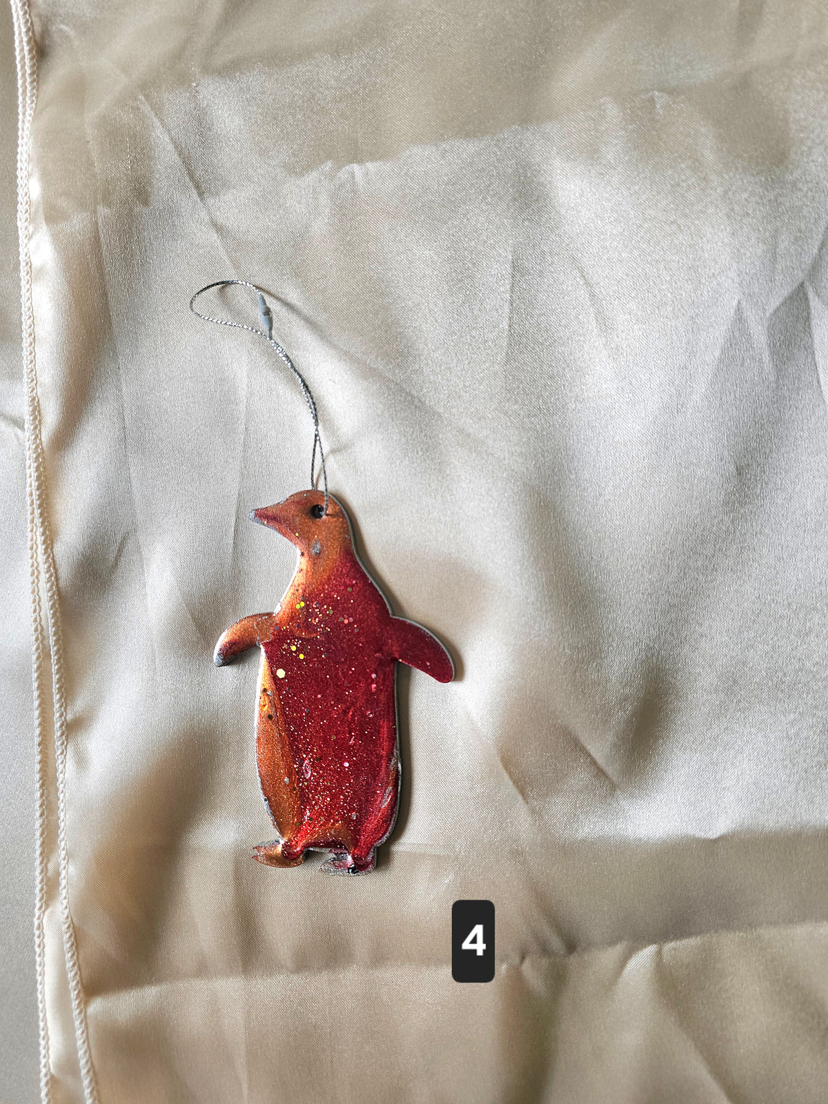 A penguin-shaped ornament or charm with a red glittery body and a silver hanging loop, placed on a light beige fabric background. A small black label with the number 4 is near the bottom.