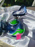 Charger l'image dans la visionneuse de la galerie, A hand-painted glass Christmas ornament with a blue velvet ribbon hanger, featuring abstract swirls of green, blue, and white. The ornament is resting on a white satin fabric surface outdoors.
