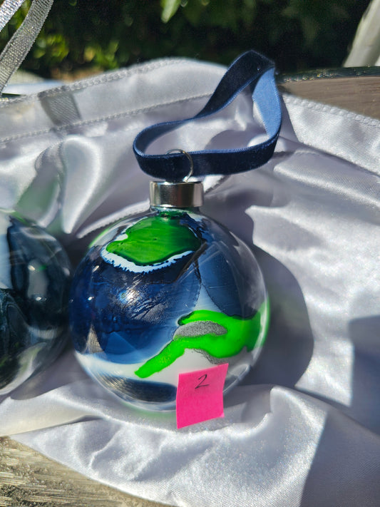 A hand-painted glass Christmas ornament with a blue velvet ribbon hanger, featuring abstract swirls of green, blue, and white. The ornament is resting on a white satin fabric surface outdoors.