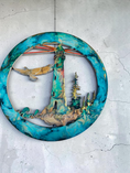 Load image into Gallery viewer, A circular abstract wall art piece featuring a lighthouse, trees, a bird, and radiating lines, with a turquoise, teal, and blue marbled border on a light gray textured wall.
