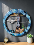 Load image into Gallery viewer, A circular resin wall art piece featuring an original design lighthouse, eagle, pine trees, and a blue marbled frame, displayed on a wooden surface against a dark gray wall with a small plant and a vintage clock nearby.
