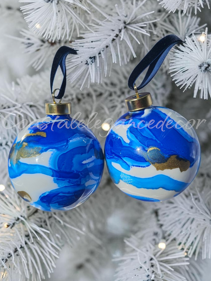 Two round blue and white marbled Christmas tree ornaments with gold caps and navy ribbons, hanging on a white artificial Christmas tree.