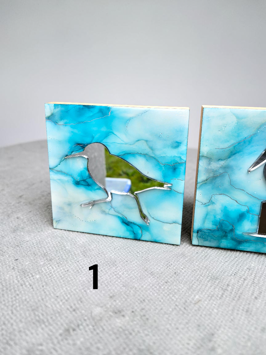Two square decorative tiles with a blue and white marbled pattern, featuring a silver metal bird cutout on the left tile, displayed on a textured gray surface.
