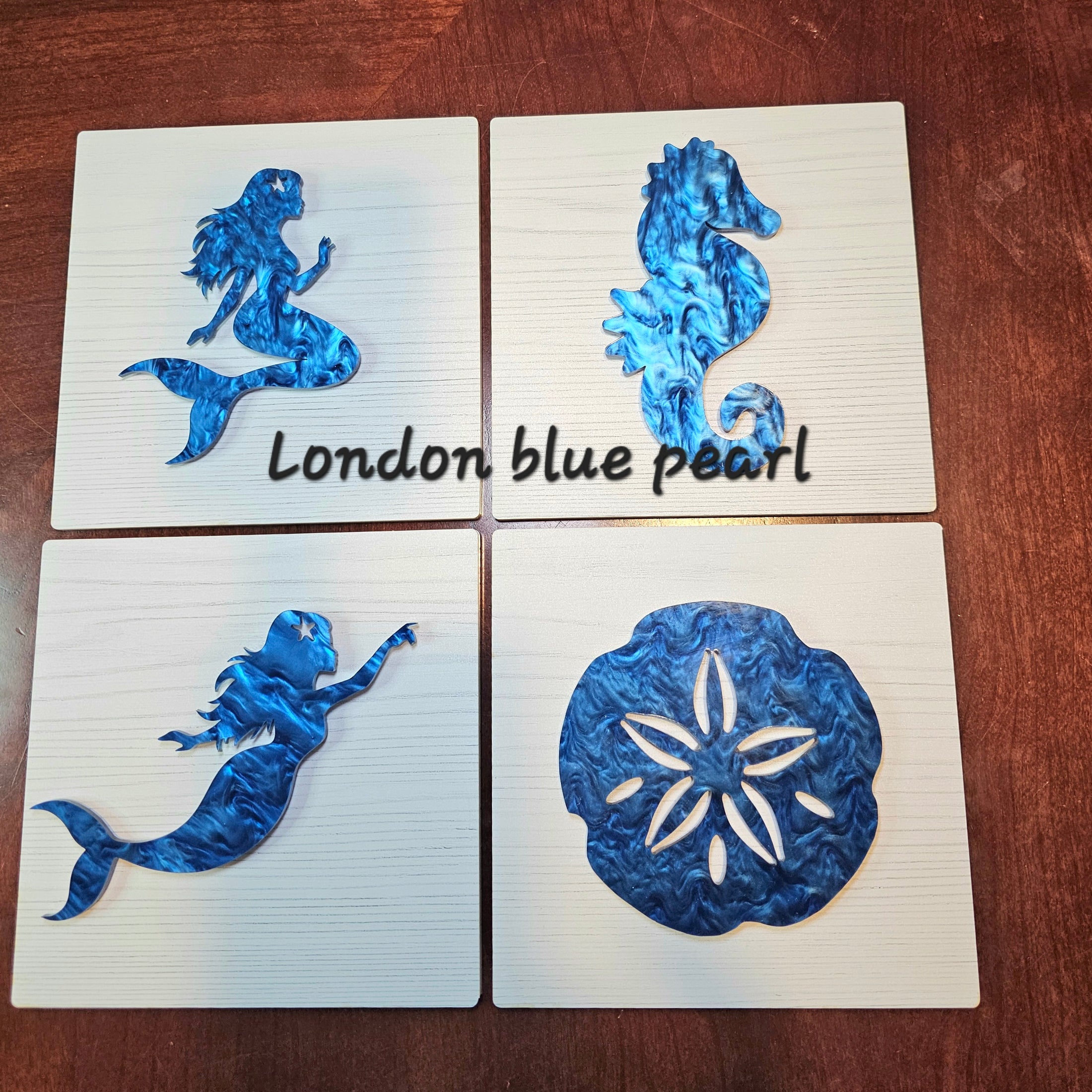 Four wall decor tiles with blue mermaid, seahorse, and sand dollar designs on a wooden surface.