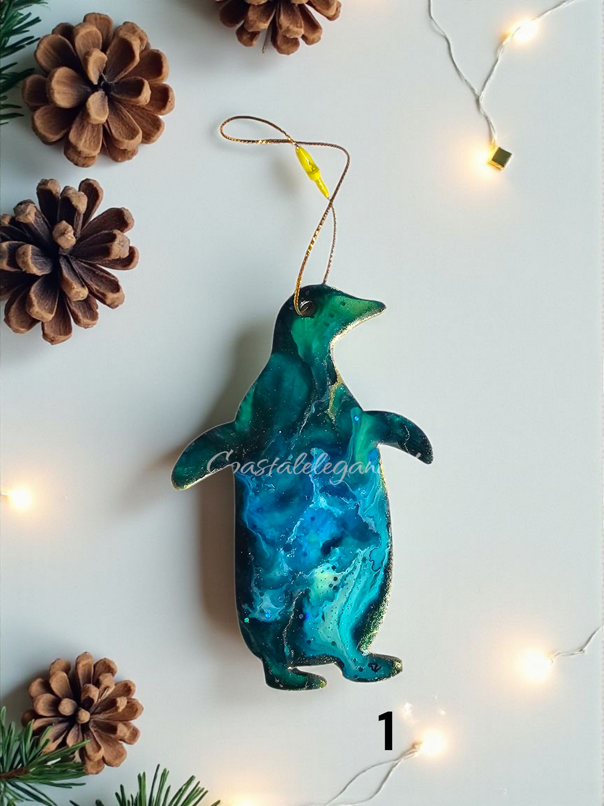 Decorative penguin resin ornament with a gradient of blue and green on a white background with pine cones and string lights.