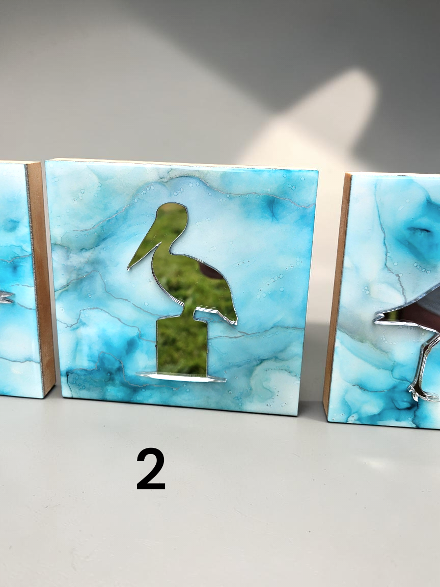 Three decorative rectangular wall art panels with blue and white marbled ink surfaces, each featuring a bird silhouette in the center. The center panel shows a mirror pelican, the right panel shows part of a shorebird, and the left panel is partially visible.
